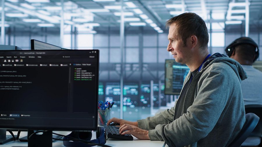 Siedzący profilem administrator IT w pracy, pisze coś na klawiaturze, obok niego ekran komputera